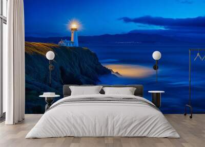 A serene lighthouse at dusk with its beacon illuminating the rocky coastline, under a tranquil blue evening sky. Wall mural