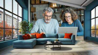 Two happy, busy middle aged professionals, man and woman, are collaborating at wooden table. They are focused on laptop, with documents and pen nearby, showcasing teamwork and productivity Wall mural