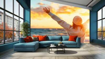 young woman enjoys beautiful sunset by sea, expressing freedom and joy with her arms raised. warm colors of sky reflect her positive emotions Wall mural