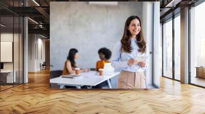 A cheerful Caucasian woman dressed in business casual attire holds a digital tablet in a contemporary office with diverse colleagues in the background. Wall mural