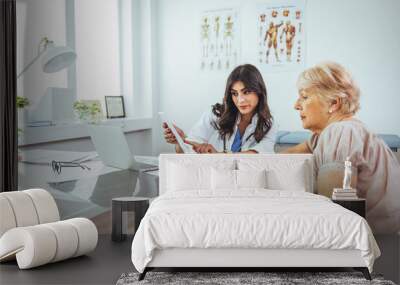 A female doctor sits at her desk and chats to an elderly female patient while looking at her test results. Patient Having Consultation With Female Doctor In Office Wall mural