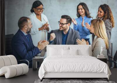 A group of diverse business professionals celebrate success, clapping for a smiling male colleague seated with a laptop in a modern office. Wall mural