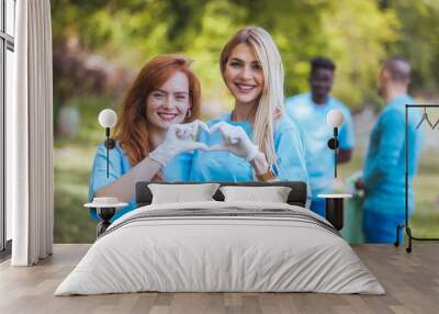 A portrait of two volunteers, standing, hugging, smiling, making a heart with their hands and looking at the camera, after cleaning the public park, have the same blue T-shirts. Wall mural