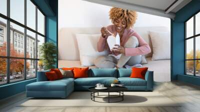 An African-American woman eats a pasta salad at home, a happy face, sits on a sofa and eats fast food that she ordered through the app. The concept of fast food and delivery Wall mural