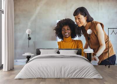 An Asian businesswoman and her Black colleague examine data on a laptop in a modern workspace, engaging in teamwork while dressed in smart casual attire, with coffee at hand. Wall mural