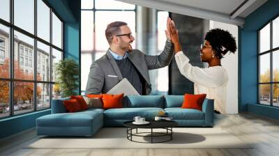 Coworkers celebrating achievement at office. Celebrating success. Happy young man standing in office and giving high five to his colleagues. Two business people high-five. Job well done Wall mural