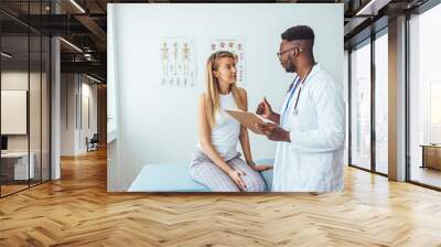 Doctor discussing treatment with cheerful smiley female patient. Happy physician and young man talking and laughing sitting on examination bed in modern clinic or hospital Wall mural