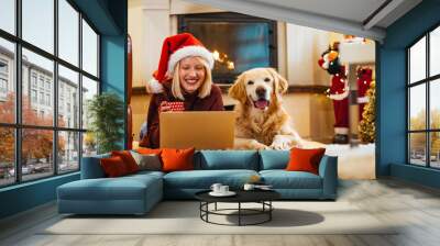 Modern young woman spending Christmas morning at home relaxing on floor with her dog in living room watching film on laptop. They just love Christmas. Wall mural