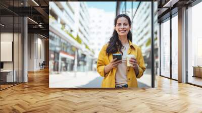 Modern young woman walking on the city street texting and holding cup of coffee. Business woman holding smartphone and looking away outdoors.  Wall mural