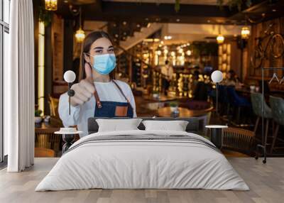 Portrait of beautiful waitress wearing protective face mask while holding touchpad and looking at the camera in a pub or restaurant during COVID-19 epidemic. Wall mural