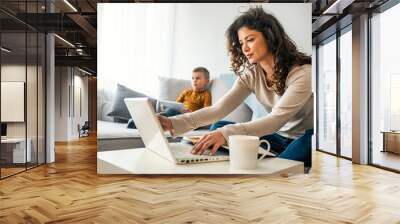 Smiling mom working at home with her child on the sofa while writing an email. Young woman working from home, while in quarantine isolation during the Covid-19 health crisis Wall mural