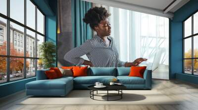 Tired african American millennial female worker sit at desk touch back suffer from lower spinal spasm, hurt unhealthy biracial woman stretch have strong backache, incorrect posture concept Wall mural