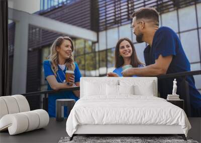 Two female and one male healthcare worker in blue scrubs share a light-hearted moment with coffee cups in hand, standing outside a medical facility. Wall mural