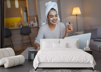 Woman with cosmetic face mask shopping online from her home. Leisure time at home. pPortrait of beautiful woman with facial taking a coffee at home while use internet with modern laptop computer Wall mural