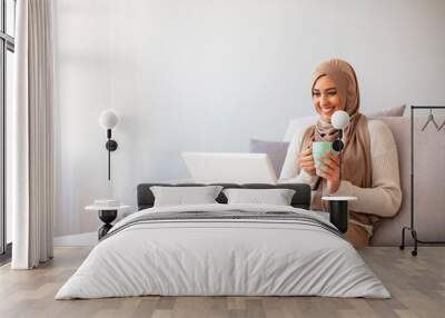 Young Arabic female entrepreneur wearing a hijab working on a laptop and drinking coffee while sitting at living room. Muslim woman with laptop at home. Wall mural