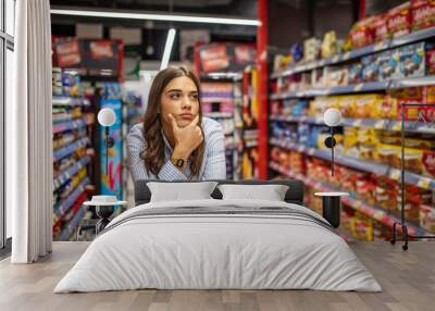 Young woman staying confused in grocery store. Confused woman doesnt know what to buy. Young woman with a shopping cart at supermarket. Looking at supermarket shelf Wall mural