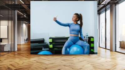 A beautiful young caucasian girl takes a selfie in the gym while sitting on a fitness ball and holding a bottle of water Wall mural