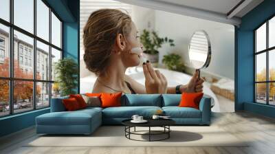 A woman in casual attire applying face skincare with a brush, holding a handheld mirror. Indoor background features natural light and green plants Wall mural