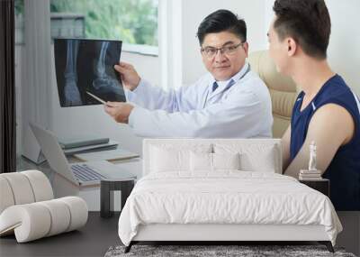 Adult Asian man in medical apparel demonstrating X-ray picture of legs to young guy while sitting at table in doctor's office Wall mural