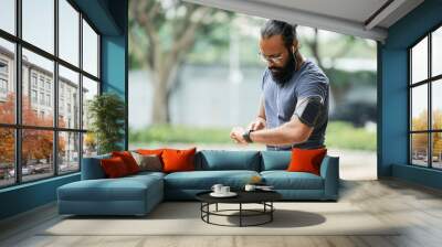 Bearded athletic Indian man in glasses checking time on smartwatch while having workout outdoors on blurred background Wall mural