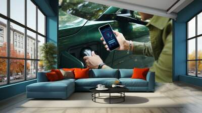 EV owner checking smart charger app on smartphone when plugging charging cable Wall mural