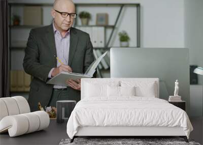 Mature businessman in suit making notes in his notebook and looking at computer monitor, he planning his work at office Wall mural