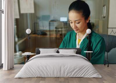 Portrait of a focused Asian female medical professional in green scrubs looking at a tablet in a modern office Wall mural