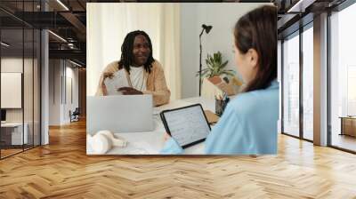 Professional meeting between two colleagues in a contemporary office setting, sharing ideas with digital tablets and laptops while surrounded by plants and office supplies Wall mural