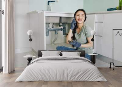 Proud cheerful young Asian woman sitting on kitchen floor and talking on phone after fixing leaking sink pipe Wall mural