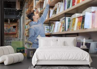 Side view of scholar boy standing before bookshelves reaching out for book  Wall mural