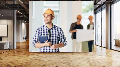 Smiling young engineer in hardhat holding tablet computer and looking around inside the house his team just finished Wall mural