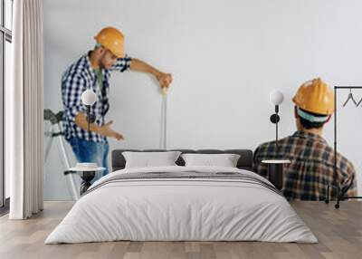 Vertical shot of professional construction worker standing on stepladder holding tape rule measuring wall length, his colleagues helping him, copy space Wall mural