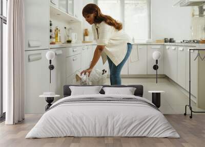 Woman in modern kitchen cleaning and throwing garbage into bin while standing near counter. Light streams through window illuminating neat and organized space Wall mural