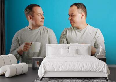 Young adult male twin siblings sitting at table looking at each other holding cups with coffee, blue background Wall mural