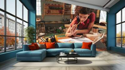 Young curly woman leaning over messy table and looking at draft with new project thoughtfully with pencil in her mouth Wall mural