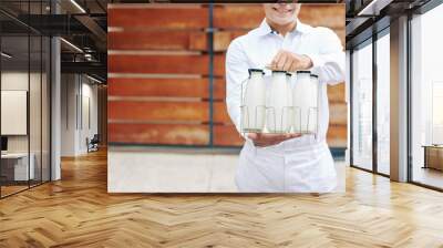 Young milkman in perfect white uniform standing against wooden fence holding glass bottles with milk, head cropped shot, copy space Wall mural