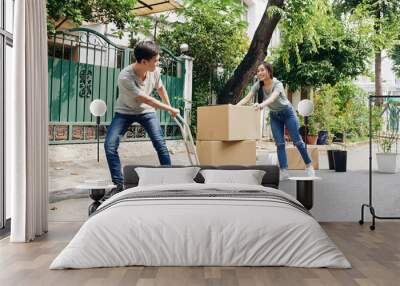 Young Vietnamese couple using trolley cart when moving in new house and transporting cardboard boxes and plants Wall mural