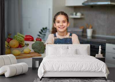 Front portrait of a little girl, in a jeans and t shirt, holding a yellow pumpkin, seated at table in the kitchen. Wall mural