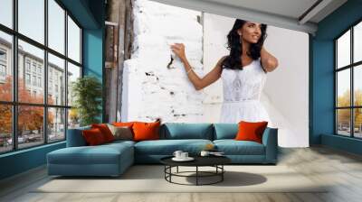 Profile portrait of a brunette splendid bride in a white wedding dress, touching her hair lightly, poses near a old white wall, looking away. Wall mural