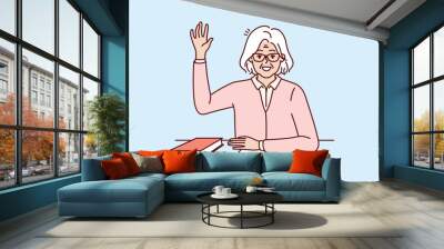 Happy elderly woman student sits at school desk with book and raises hand up wanting to ask question to teacher. Viable positive pensioner student getting education after retirement Wall mural