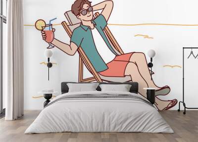 Man tourist is sunbathing on beach sitting in sun lounger and drinking fruity refreshing cocktail Wall mural