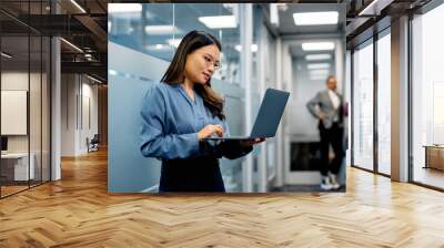 Asian businesswoman using laptop in hallway of office building. Wall mural