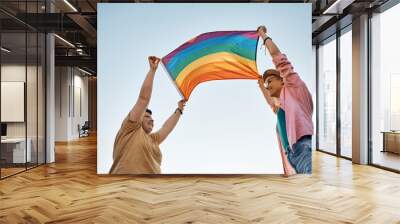 Below view of happy lesbian couple holding rainbow flag against the sky. Wall mural