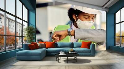 Black civil engineer with face mask sneezing into elbow outdoors. Wall mural