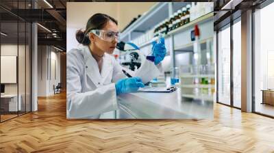 Female scientist taking notes while analyzing blue chemical liquid in lab. Wall mural