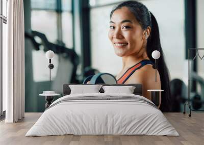 Happy Asian athletic woman lifts hand weights during sports training at gym and looks at camera. Wall mural