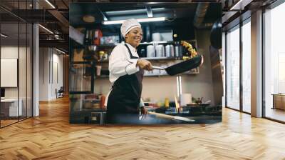 Happy black female chef has fun while preparing food in frying pan at restaurant kitchen. Wall mural