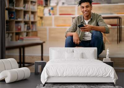 Happy black student using mobile phone at university library and looking at camera. Wall mural