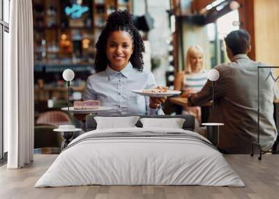 Happy black waitress serving dessert in a cafe and looking at camera. Wall mural