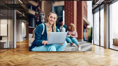 Happy female college student e-learning on laptop at campus and looking at camera. Wall mural